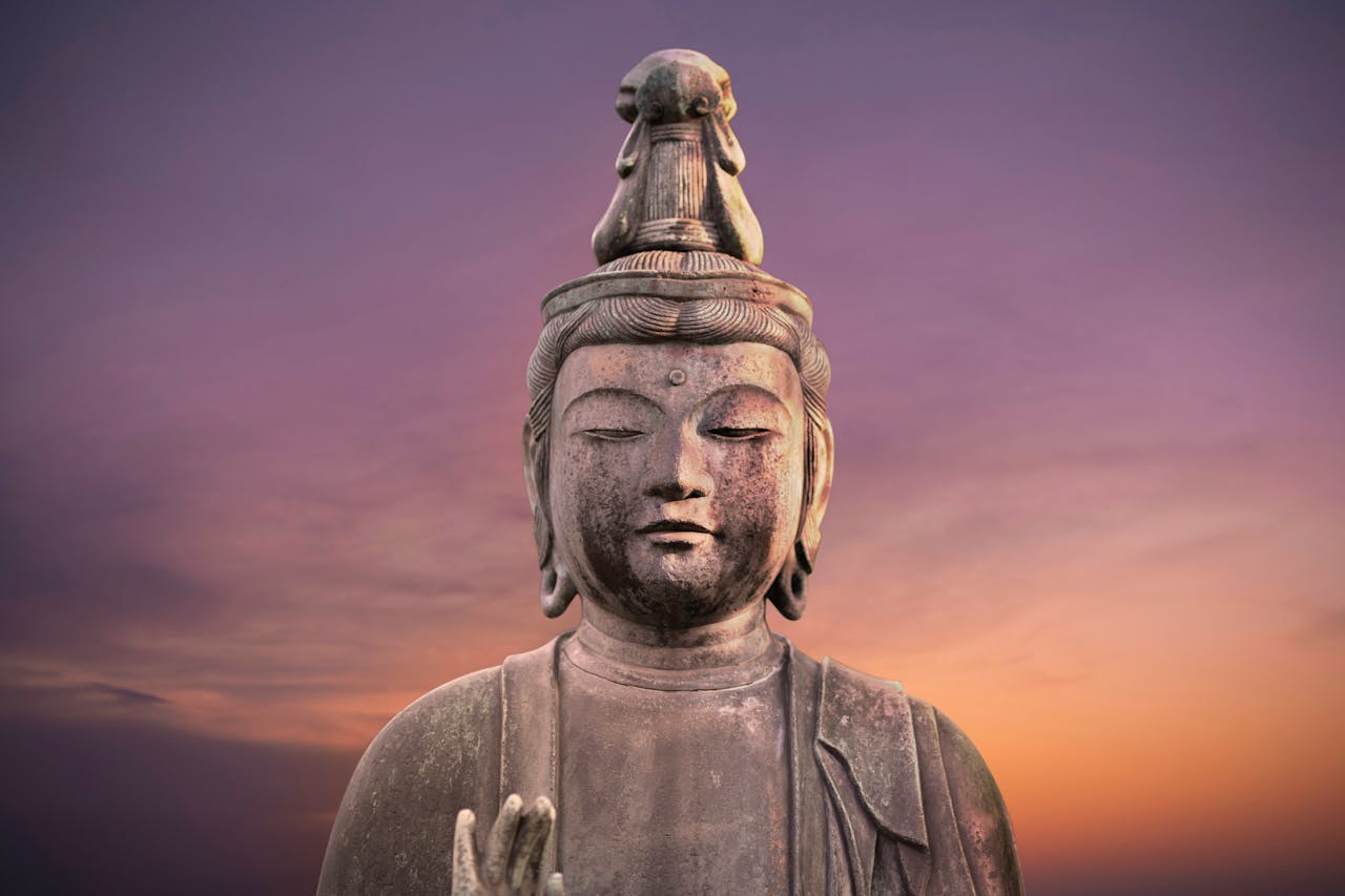 The image shows a stone Buddha statue with a serene facial expression and an elaborate headdress, set against a purple and orange sunset sky. The statue displays the traditional meditative pose, embodying the calm and introspective nature of Buddhist philosophy. This ancient sculpture represents core Buddhist principles of mindfulness and inner peace, making it an ideal visual complement to an article about Buddhism's teachings.
